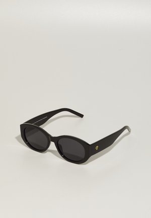 Black sunglasses with oval lenses, thick plastic frames, and a shiny gold logo on the temple. Lenses are dark and slightly reflective.