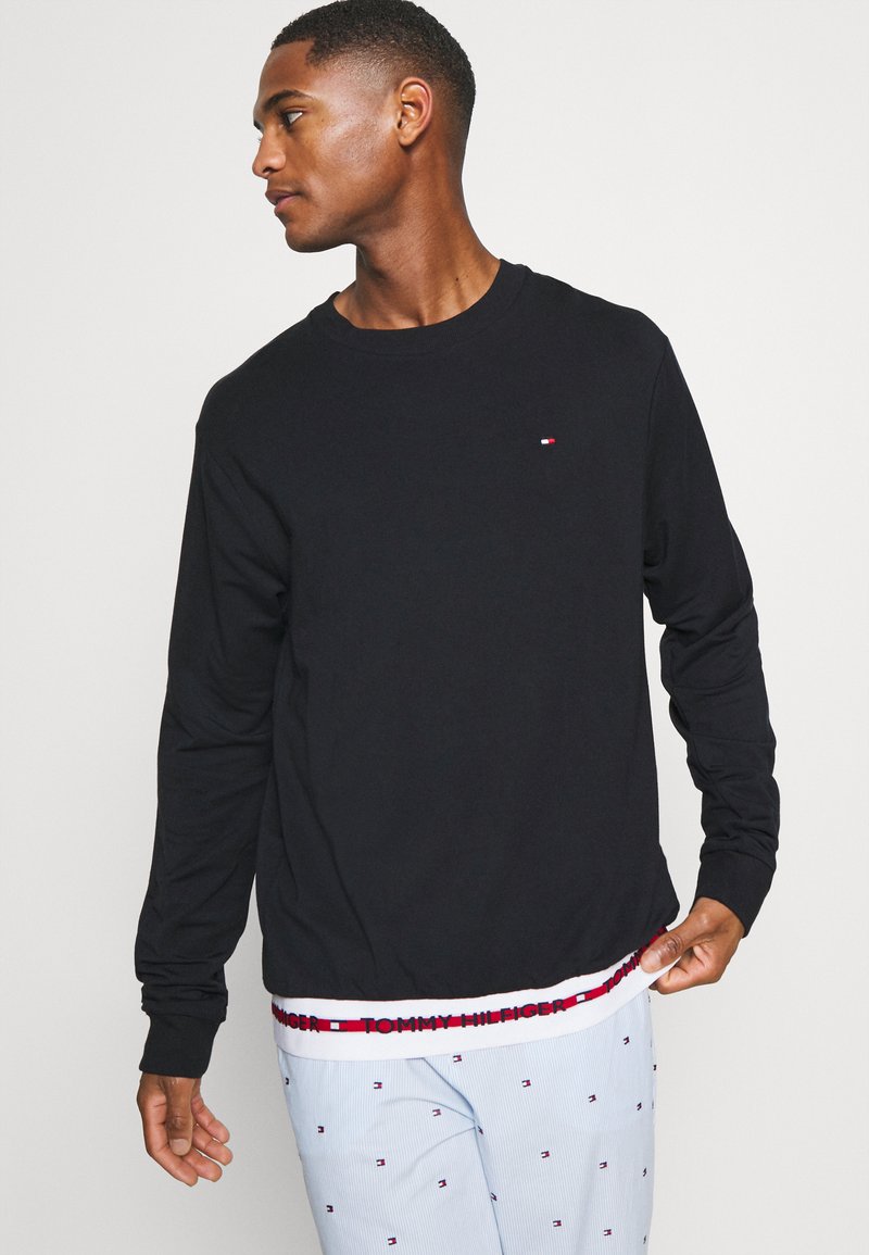 Uomo in camicia nera a maniche lunghe con piccolo logo, tirando l'orlo per mostrare una banda bianca con il testo "Tommy Hilfiger", e pantaloni azzurri chiari con piccoli loghi.