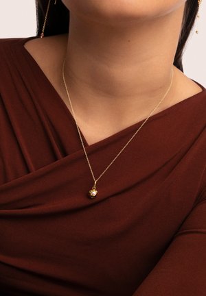 Collier en or avec un pendentif rond, présentant des détails floraux, reposant sur un haut en tissu marron, mettant en valeur la longueur de sa chaîne et son design délicat.