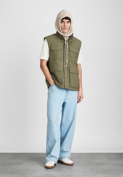 Olivno zelena brezrokavnik s štirimi sprednjimi žepi, nošen preko svetlega T-majice, v kombinaciji s svetlo modrimi širokimi hlačami in belimi supergami.