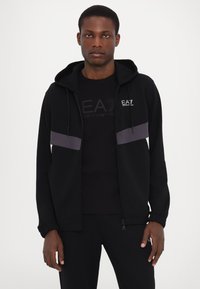 Black zip-up hoodie with grey accents, drawstring hood, and ribbed cuffs. Worn over a black t-shirt featuring a logo design.