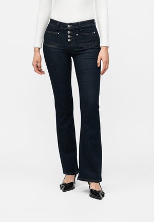 Kvinde iført mørkeblå jeans med høj talje og bootcut, fire knapper, forlommer, en hvid top med lange ærmer og sorte spidse hæle.