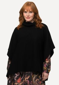 Mustapaino poncho, jossa on korkea kaulus, rento istuvuus ja sileä rakenne, yhdistettynä värikkääseen kuviolliseen mekkoon alapuolella.