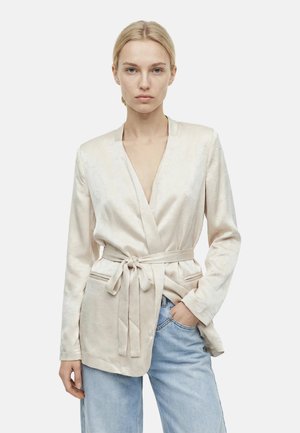 Donna bionda con blazer color crema con cintura e scollo a V profondo, jeans azzurri chiari, una mano in tasca, sfondo neutro.