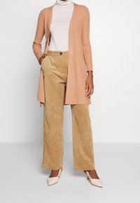 Beige gerippter langer Cardigan über einem hellen Rollkragenpullover, kombiniert mit weiten Cordhosen und kremfarbenen spitzen Absätzen.