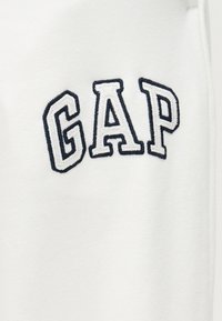 Άσπρο ύφασμα με κεντημένα γράμματα "GAP" με μαύρο περίγραμμα σε στυλ κολεγιακής γραμματοσειράς, τοποθετημένα στο κέντρο του υφάσματος.