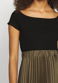 Musta ribattuva paita lyhyillä hihoilla siirtyy khakinväriseen liesituulettimen hameeseen, jossa on joustava vyötärö, korostaen rakennetta ja väri-eroa.