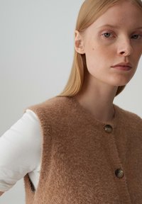 Gilet di maglia marrone morbido con scollo rotondo e tre bottoni, indossato sopra una camicia bianca a maniche lunghe. Texture morbida, colore caldo.