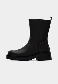 Μαύρη δερμάτινη μπότα μέχρι τη μέση της γάμπας με παχιά σόλα και χαμηλό τετράγωνο τακούνι, που διαθέτει τρ άβηγμα στο πίσω μέρος και λείος στρογγυλεμένος νύχιος.