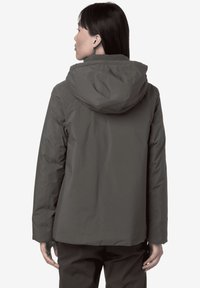 Veste imperméable grise avec capuche, présentant une texture lisse et une coupe droite, conçue pour une utilisation en extérieur. Accents réfléchissants visibles.