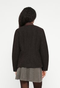 Vrouw met lang, donker haar, gekleed in een donkere, gestructureerde jas over een grijze rok, staande met haar rug naar de kijker tegen een effen lichte achtergrond.