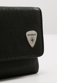 Portefeuille en cuir noir avec surface texturée, quincaillerie argentée présentant un logo triangulaire, et couture visible le long des bords.
