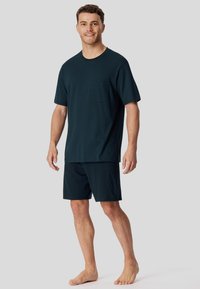 Un joven de pie descalzo, vistiendo una camisa de manga corta con un estampado oscuro y pantalones cortos oscuros a juego, frente a un fondo sencillo.