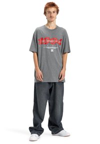 Jeune personne aux cheveux roux portant un t-shirt graphique gris surdimensionné, un jean sombre ample et des baskets blanches, se tenant devant un fond blanc.