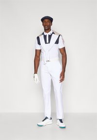 Biała koszulka polo golfowa z granatowymi akcentami, zestawiona z białymi spodniami i butami golfowymi. Lekki sweter zarzucony na ramionach, biała rękawiczka.