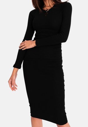 Femme portant une robe midi noire cintrée à manches longues, avec des colliers dorés superposés et un pendentif en forme de croix, debout avec un bras croisé.