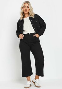 Giacca in denim nero con chiusura a bottoni, abbinata a pantaloni neri a vita alta. Il modello indossa un top chiaro e sneakers bianche con dettagli colorati.