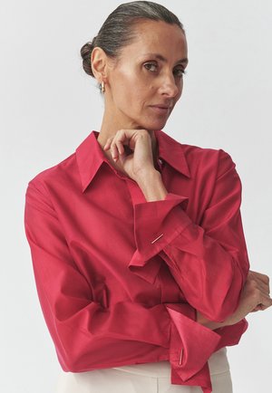 Blusa rossa a maniche lunghe con colletto strutturato e polsini arrotolati, caratterizzata da accenti hardware minimalisti in argento. Texture del tessuto liscia.