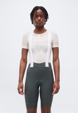 Mujer con una camisa beige de manga corta y pantalones cortos de ciclismo verdes oscuros con tirantes blancos, de pie contra un fondo claro y liso.