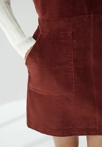 Rotes Cord-Kleid mit vertikalen Streifen und einer Vordertasche, kombiniert mit einem strukturierten weißen Langarmshirt.