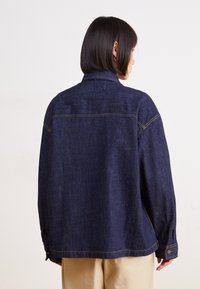 Chaqueta de mezclilla en azul oscuro, con cuello tipo solapa, mangas largas y detalles de botones en los puños. Costuras visibles a lo largo de las costuras.