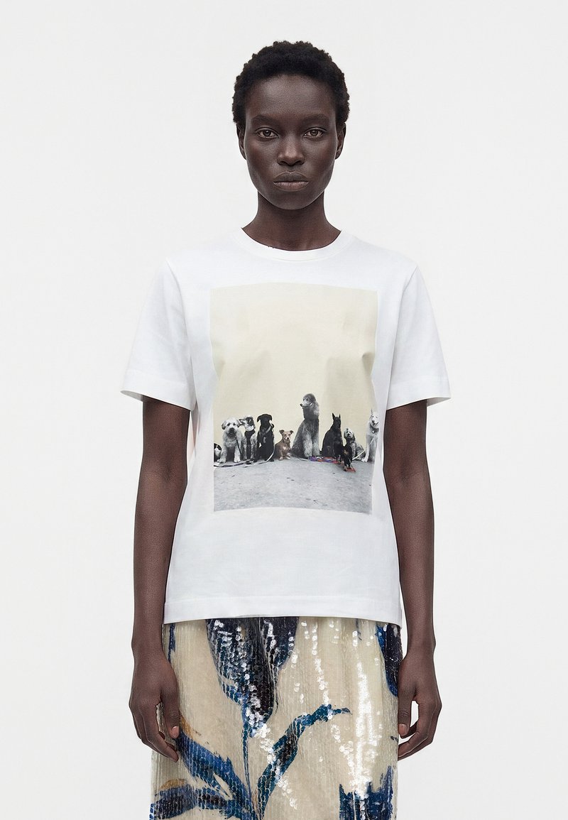 Femme portant un T-shirt blanc avec l'impression de huit races de chiens différentes assises sur le trottoir, associée à une jupe fleurie brillante.