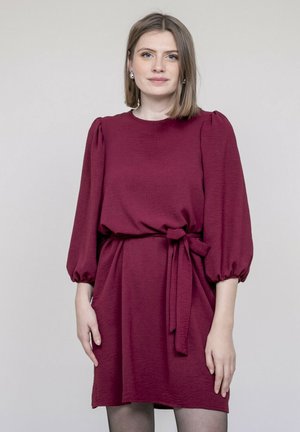 Robe bordeaux avec manches ballon, col rond et ceinture à nouer à la taille. Le tissu a une texture apparente et tombe juste au-dessus du genou.