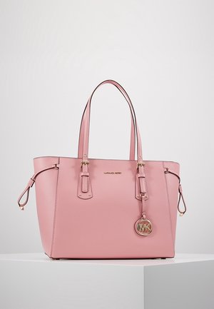 Pink leather tote bag with double handles, Michael Kors logo, and round MK charm hanging from one handle, placed on white pedestal against gray background.