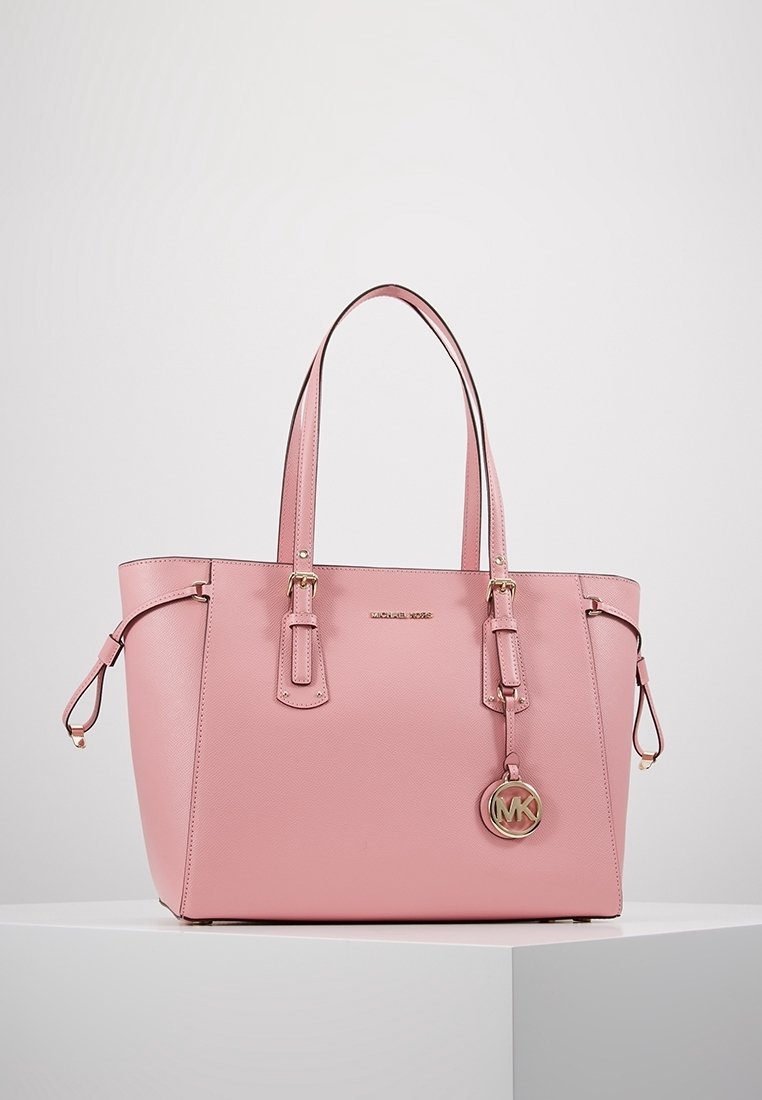 Sac cabas en cuir rose avec double poignées, logo Michael Kors et pendentif rond MK suspendu à une poignée, posé sur un socle blanc sur fond gris.