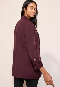 Burgundy zakó, testhez álló szabással, kétsoros hajtókákkal, gombos mandzsettákkal és diszkrét oldalsó zsebekkel, fekete slim-fit nadrággal párosítva.