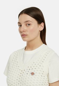 Gilet bianco lavorato a maglia con scollatura a V, caratterizzato da un motivo testurizzato e un piccolo logo Dickies. Indossato sopra una t-shirt bianca.
