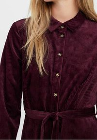 Donna che indossa un vestito di velluto a coste bordeaux con maniche lunghe, bottonato e con colletto e cintura in vita, con lunghi capelli biondi che scendono su una spalla.