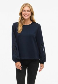 Giovane donna che sorride, indossando un top a maniche lunghe blu navy con motivi floreali intagliati sulle maniche e jeans neri.