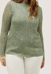 Pull en maille vert clair à manches longues, présentant un motif ajouré. Col et poignets soulignés, assortis à un pantalon blanc.