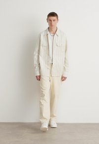 Chemise à boutons de couleur claire avec des rayures verticales, associée à un pantalon crème et des baskets blanches. Design simple et coupe décontractée.