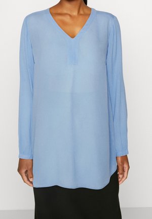 Kvinde iført en lyseblå langærmet V-hals bluse og et sort nederdel, stående mod en ensartet baggrund.