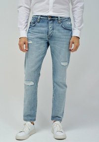 Vaqueros destrozados de color azul claro con desgastes en las rodillas, cinco bolsillos y un corte recto, llevados con zapatillas blancas y una camisa blanca de botones.