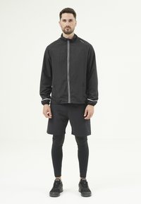 Svart lättviktsjacka med vit dragkedja och reflexdetaljer, i kombination med svarta shorts och leggings. Atletiska skor fulländar outfiten.