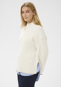 Femme blonde dans un pull en maille blanche superposé sur une chemise rayée bleue, regardant légèrement sur le côté devant un fond blanc uni.