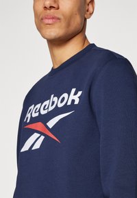 Muškarac nosi tamnoplavi Reebok duksericu s bijelim i crvenim logotipom, izrezani pogled koji prikazuje vrat, bradu i gornji dio torsija.