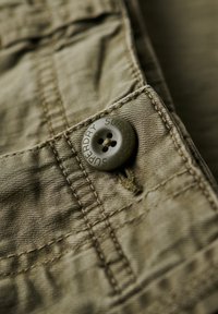 Tejido caqui con una superficie texturizada, que presenta un botón redondo marrón grabado con "SUPERDRY" y costuras reforzadas a lo largo de los bordes.