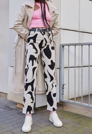 Personne portant un trench beige, un haut rose sans manches, un pantalon à motif abstrait noir et blanc et des baskets blanches, debout près d'une rambarde métallique.