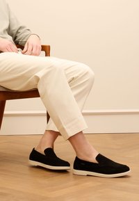 Black suede slip-on shoes with a rounded toe, white rubber sole, and minimal design. Worn with light-colored cropped trousers.