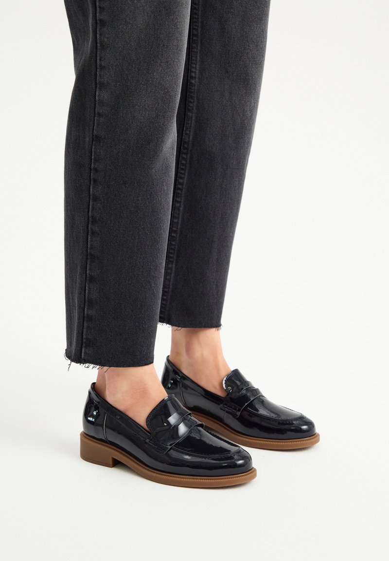 Black patent leather loafers with a low brown heel, featuring a silver accent on the front, paired with black cropped jeans.