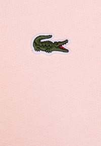 Tissu en coton rose avec un logo de crocodile brodé en vert et rouge, bordé de blanc, mettant en valeur une surface en maille texturée.