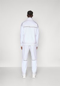 White tracksuit with a zip-up jacket and elastic waistband pants. Features textured fabric, gold accents, and fitted cuffs.