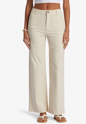 Femme portant un pantalon crème taille haute à jambes larges, un top court blanc, un bracelet coquillage et des sandales blanches tressées, debout les mains relâchées le long du corps.