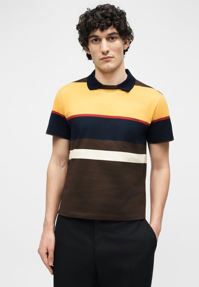 Young man with curly hair wearing a short-sleeve polo shirt with yellow, navy, brown, white, and red horizontal stripes.