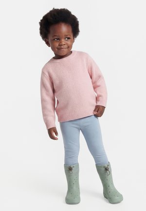 Junges Kind mit lockigem Haar, trägt einen pinken Pullover, hellblaue Hose und grüne Gummistiefel, steht leicht geneigt und lächelt.