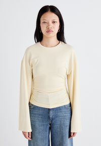 Top lungo a maniche lunghe di colore giallo chiaro con scollo rotondo e dettagli arricciati in vita. Tessuto morbido e liscio. Indossato con jeans a gamba ampia blu.